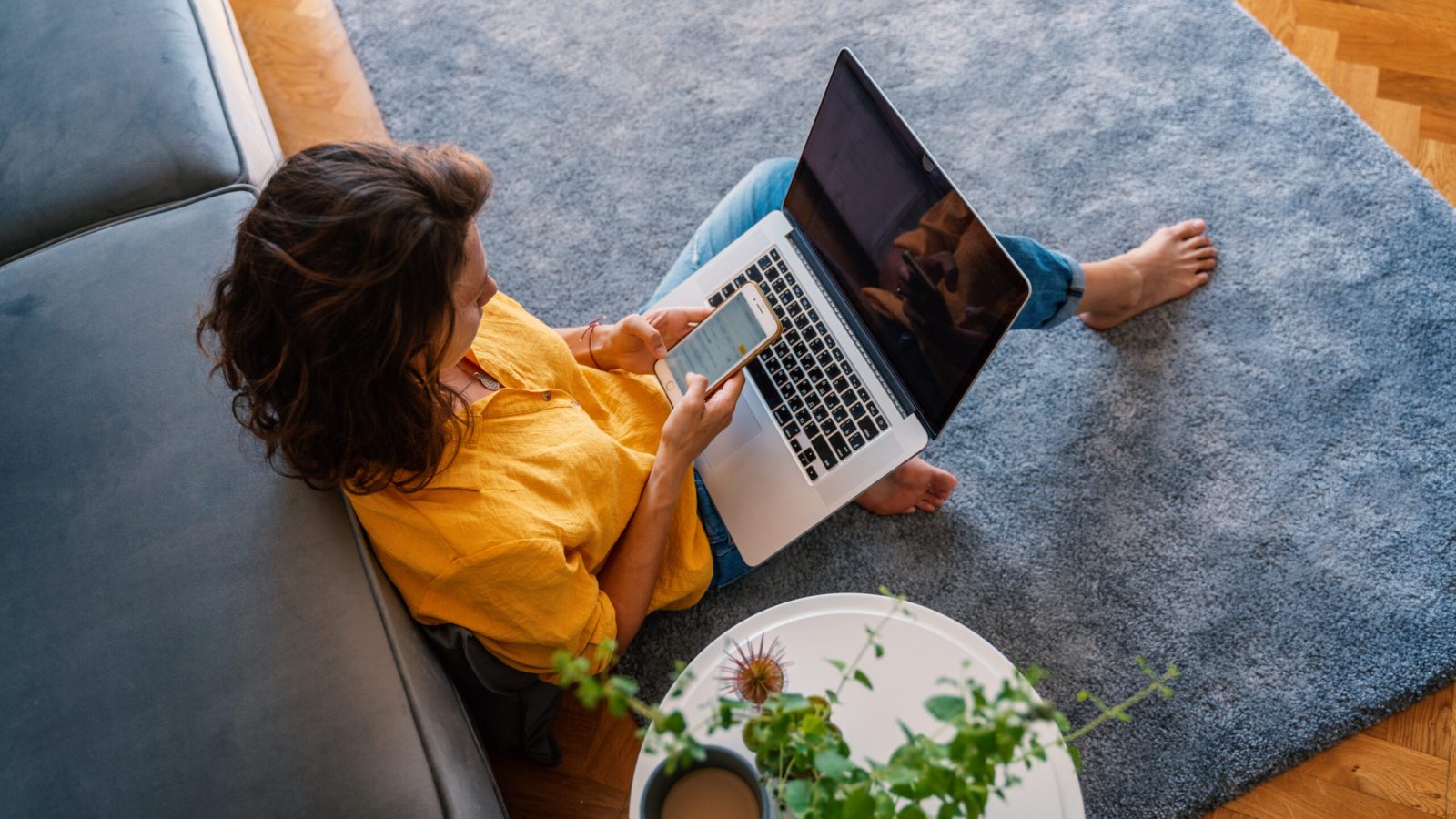 young girl in a yellow shirt woman working on a laptop at home using a smartphone in the living room on the carpet, distant work and education.